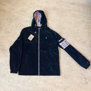 Burberry Windbreaker  - Svart vindjacka från Burberry med klassiskt rutigt foder i huvan och på ena ärmen. Jackan har dragkedja framtill, snörning i huvan och Burberry-logga på bröstet samt ärmen. Tillverkad i glansig polyester och har en loose passform.