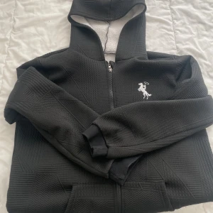 Svart hoodie med dragkedja och mönster - Svart hoodie med dragkedja och huva, fodrad med mjukt vitt tyg på insidan. Tröjan har ett diskret geometriskt mönster och en broderad vit häst på bröstet. Perfekt för kyliga dagar och riktigt skön att ha på sig.