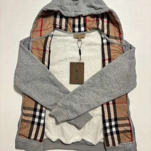 Grå Burberry hoodie - Grå hoodie från Burberry, 10/10 kvalité med klassiskt rutigt foder på insidan av huvan och längs öppningen. Tröjan har hel dragkedja, snörning i huvan och broderad logga på bröstet. Perfekt för de kommande kyliga dagar. Storleken är M, skickas inom 24 timmar. 