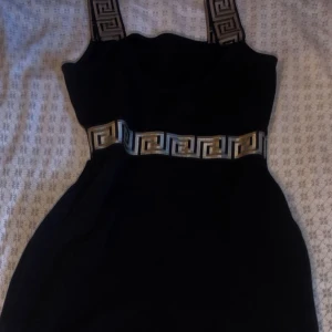 Svart kortklänning med mönstrade band - Svart kortklänning med breda axelband och ett snyggt geometriskt mönster i guld och silver längs midjan och banden. Klänningen har en figurnära passform och ärmlös design, perfekt för dig som gillar stilrena detaljer med lite edge.