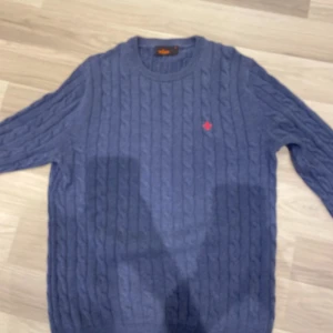 Blå kabelstickad tröja Morris - Kabelstickad blå tröja från Morris med rund hals och ett rött broderat emblem på bröstet. Tröjan har lång ärm och ribbade muddar vid ärmslut och nederkant. Perfekt för dig som gillar klassisk och stilren look.