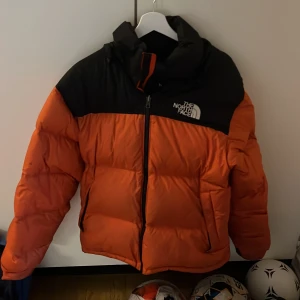 Orange dunjacka från The North Face - Säljer en fet orange och svart dunjacka från The North Face. Jackan har hög krage, huva och dragkedja framtill. Den är riktigt puffig och har logga på både bröstet och ryggen. Perfekt för kalla dagar när du vill hålla stilen och värmen.