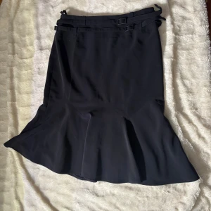 Mörkblå kjol med spännen från Orwell - Stilren mörkblå kjol från Orwell med två dekorativa spännen i midjan. Kjolen har en lätt utsvängd form nertill och är tillverkad i ett mjukt polyestertyg som ger ett snyggt fall. Perfekt för dig som gillar minimalistisk och elegant stil. Orwell är ett fint märke med bra kvalitet som är populär i Tyskland. Har inte sett Orwell i Sverige alls. Köpt i Frankfurt am main. Markerad storlek 36. Har 38 cm * 2 i midjan. 61 cm lång . Skick 10/10.