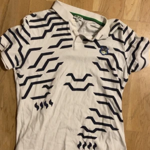 Vit mönstrad Kenzo piké, storlek M - Säljer en vit pikétröja från Kenzo med svart grafiskt mönster över hela tröjan och klassisk krage. Tröjan har korta ärmar, knappar vid halsen och en broderad Kenzo-logga på bröstet. Perfekt för dig som gillar statement-plagg med cool design.