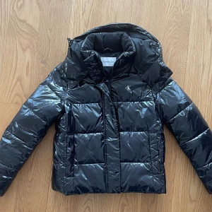 Svart pufferjacka Calvin Klein Jeans - Svart glansig pufferjacka från Calvin Klein Jeans med huva och dragkedja. Jackan har CK-logga på bröstet och stor logotyp på insidan. Fylld med syntetmaterial och har långa ärmar. Perfekt för kalla dagar och har en modern, boxig passform.