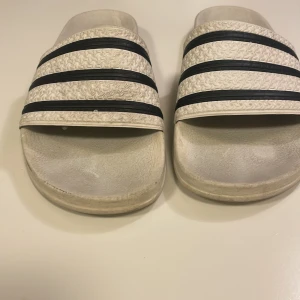 Adidas vita sandaler med svarta ränder - Klassiska Adidas sandaler i vitt med tre svarta ränder över foten. Tillverkade i syntetmaterial med präglad struktur på remmen och Adidas-logga på sidan. Perfekta för sommardagar eller att chilla i hemma.