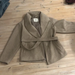 Beige kappa från ONLY med bälte - Snygg beige kappa från ONLY med bred krage och knytskärp i midjan. Jackan har två fickor framtill och är fodrad på insidan. Perfekt för dig som vill ha en stilren och trendig look under kyligare dagar.