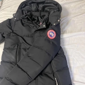 Svart dunjacka från Canada Goose M - Säljer en svart dunjacka från Canada Goose i storlek M. Jackan har huva, dragkedja och tryckknappar framtill samt två stora fickor. Klassisk Canada Goose-logga på ärmen och diskret logotyp vid fickan. Perfekt för kalla vinterdagar.