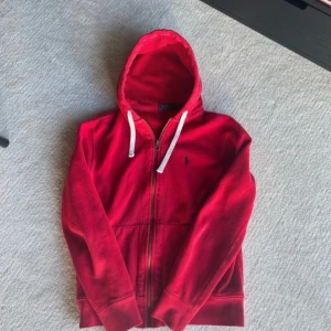 Röd hoodie från Polo Ralph Lauren - Riktigt snygg röd hoodie från Polo Ralph Lauren med dragkedja och vit snörning i huvan. Klassisk broderad logga på bröstet och mjukt material som känns skönt mot huden. Perfekt för lager-på-lager och chill dagar.