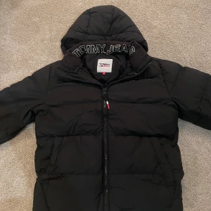Svart pufferjacka från Tommy Jeans XS - Svart pufferjacka från Tommy Jeans med huva och broderad logga på insidan av kragen. Jackan har dragkedja framtill, sidofickor med tryckknapp och klassisk pufferdesign. Perfekt för kalla dagar och riktigt snygg streetstil. Inga defekter och väldigt varm.
