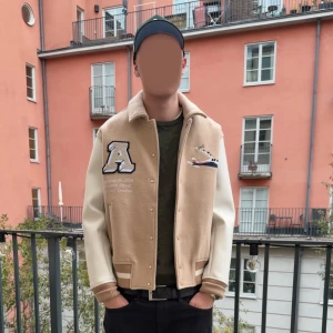 Beige bomberjacka Axel Arigato - Säljer en beige och vit bomberjacka från Axel Arigato med stora broderade bokstäver på ryggen och detaljer som ett flygplan och ett stort A på bröstet. Jackan har ribbade muddar, knappar framtill och kontrastfärgade ärmar i syntetmaterial.