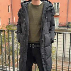 Svart lång pufferjacka med huva - Säljer en svart lång pufferjacka med huva och dragkedja framtill. Jackan har quiltad design, två sidofickor och är långärmad. Perfekt för kalla dagar när du vill hålla dig varm och ändå se chill ut. Passar dig som gillar en oversized vibe.