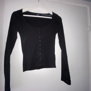Svart långärmad topp med knappar - Svart långärmad topp från Bik Bok med rundad halsringning och knappar längs hela framsidan. Modellen är slim, perfekt för att styla med låga jeans. Mjukt material som sitter skönt mot kroppen.