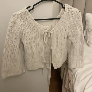 Vit stickad kofta med knytband - Superfin vit/beige stickad kofta med långa ärmar och tre knytband framtill. Koftan har en lätt croppad passform och är perfekt att slänga över ett linne. Materialet är mjukt och luftigt, vilket gör den skön att bära under både vår och sommar. 