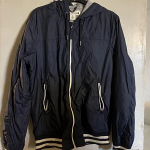 Mörkblå vindjacka från L.O.G.G. - Snygg mörkblå vindjacka från L.O.G.G. med huva och vita detaljer på dragkedja och fickor. Jackan har ribbade muddar med vita ränder och snörning vid huvan. Perfekt för dig som gillar sportig och clean stil.