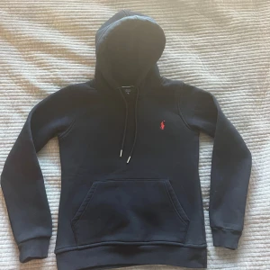 Ralph lauren hoddie  - Mörk blå hoodie från Ralph Lauren med klassisk röd logga broderad på bröstet. Tröjan har huva med snörning, magficka och ribbade muddar. Storleken är S men den är xs eftersom den sitter ganska liten på mig och jag har s. Skriv vid frågor !