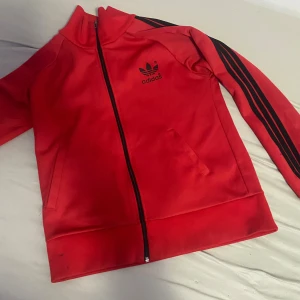 Röd Adidas ziptröja med svarta ränder - Röd Adidas ziptröja med svarta klassiska ränder längs ärmarna och stor Adidas-logga på ryggen. Tröjan har hög krage, dragkedja framtill och två fickor. Perfekt för en sportig och avslappnad stil.