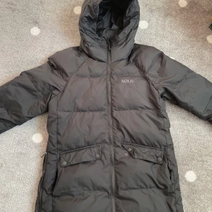 Lätt och varm pufferjacka från NOUN - Svart pufferjacka från NOUN med huva och dragkedja framtill. Jackan är lång, har två stora fickor med lock och knappar samt en hög krage som skyddar mot kyla. Perfekt för kalla dagar och har en clean, stilren look.