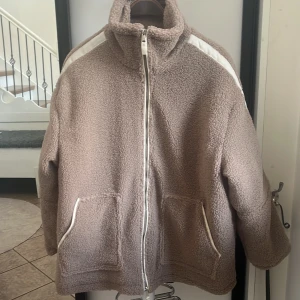 Beige teddyjacka med vita detaljer - Mysig beige teddyjacka med vita ränder längs axlarna och vita detaljer vid fickorna. Jackan har hög krage, hel dragkedja framtill och två stora fickor. Insidan är quiltad för extra komfort. Perfekt för kyliga dagar och en chill look.