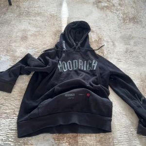Svart hoodie från Hoodrich med tryck - Svart hoodie från Hoodrich med stor logga framtill och text på ryggen. Tröjan har luva med tryck på ena ärmen. Mjuk och skön, perfekt för chill eller streetwear. 