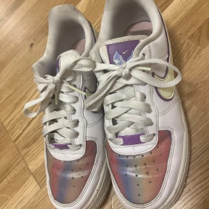 Nike Air Force 1 med regnbågstoe - Nike Air Force 1 sneakers i vitt skinn med regnbågsfärgad tå och lila detaljer. Swoosh-loggan är ljusgul med lila kant, och skorna har klassisk snörning och platt sula. 
