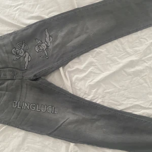 Grå jeans med strassdetaljer BoohooMAN - Säljer ett par grå jeans från BoohooMAN med snygga strassdetaljer i form av änglar och texten 'BLINGLUCI' på benen. Jeansen har klassisk femficksdesign och är tillverkade i bomull. Perfekt för dig som gillar att sticka ut med coola detaljer.