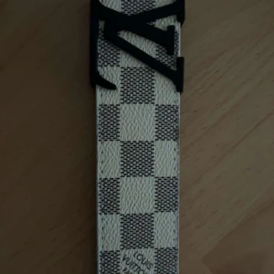 Beige rutigt skärp i skinn - Snyggt -bälte i klassiskt rutigt mönster med beige och vita toner. Bältet har en svart -logga som spänne och är gjort i skinn. Perfekt accessoar för att lyfta din outfit med en exklusiv touch.