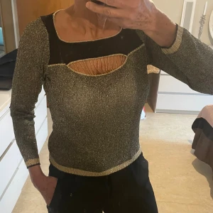 Glittrig långärmad topp från TeDu - Säljer en glittrig svart och guld topp från TeDu   coolt cut-out parti framtill. Toppen har guldfärgade detaljer vid hals, ärmslut och nederkant samt ett transparent svart parti upptill. Tyget är töjbar .det finns ingen storlek men,räknas som en smal eller en liten medium.jag har medium så den passar mig som det finns i bild 