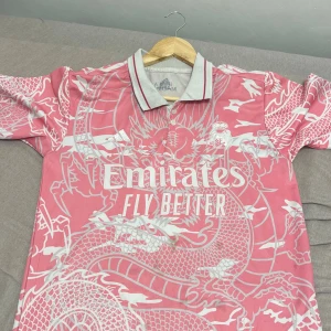 Rosa Real Madrid fotbollströja Adidas - Säljer en unik Real Madrid fotbollströja från Adidas i rosa med vit drakmönster. Tröjan har korta ärmar, krage med ränder och tryckta loggor på bröstet. Materialet är lätt och andas, perfekt för match eller träning. Emirates sponsortryck på framsidan.