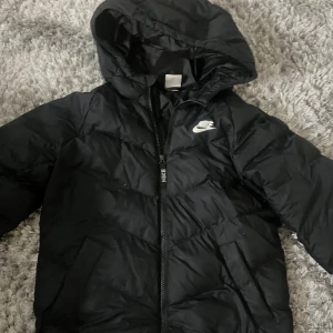 Svart pufferjacka från Nike - Svart pufferjacka från Nike med huva och dragkedja framtill. Super snygg på. Kontakta mig för minsta fundering💕