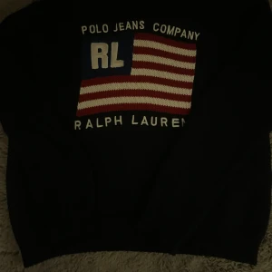 Mörkblå stickad tröja Ralph Lauren - Säljer en mörkblå stickad tröja från Ralph Lauren Polo Jeans Company med amerikansk flagga och RL-brodyr på bröstet. Tröjan är i 100% bomull och har långa ärmar samt ribbade muddar. Perfekt för dig som gillar klassisk och sportig stil.
