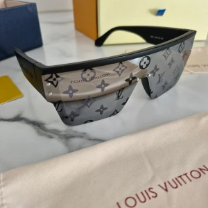 Louis Vuitton svarta shield-glasögon - Säljer ett par svarta shield-glasögon från Louis Vuitton med breda skalmar och LV-monogram över hela linsen. Glasögonen har en futuristisk, kantig form och är tillverkade i syntetmaterial. Kommer med originalfodral, ask och certifikat.