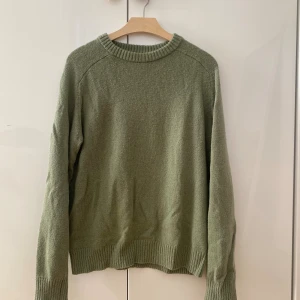 Olivgrön stickad tröja med rund hals - En olivgrön stickad ulltröja med rund halsringning och ribbade muddar vid ärmslut och nederkant. Tröjan har en avslappnad passform. Storlek S, men sitter som en M. Väl omhändertagen.