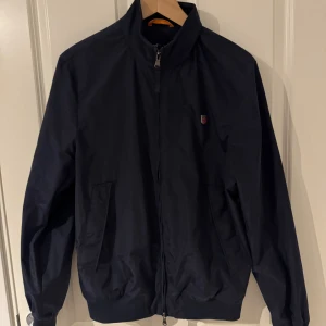 Marinblå vindjacka från Morris S - Snygg marinblå vindjacka från Morris med klassisk ståkrage, dragkedja och två snedställda fickor framtill. Jackan har diskret logga på bröstet och ribbade muddar vid ärmslut och midja. Perfekt för dig som gillar stilren och enkel design. Pris ej fast kom med frågor