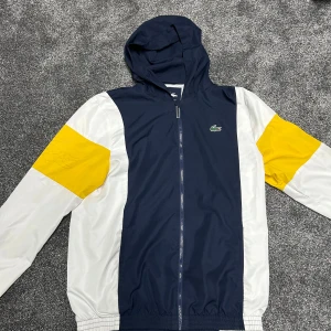 Lacoste Sport vindjacka/Tröja med huva M - Snygg vindjacka/tröja från Lacoste Sport i marinblått, vitt och gult. Jackan har huva, hel dragkedja och elastiska muddar. Ikoniska krokodilloggan på bröstet. Perfekt för dig som gillar sportig och stilren look.