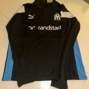 Svart Puma Marseille half zip träningströja - Svart långärmad träningströja från Puma med Olympique de Marseille-logga och vit rand på axlarna. Half zip framtill, tryckta loggor och blå detaljer på sidorna. Perfekt för fotbollsträning eller chill. Materialet är polyester.