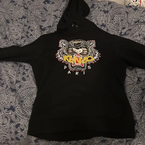 Svart Kenzo hoodie med tigertryck - Svart hoodie från Kenzo med ikoniskt broderat tigerhuvud och färgglad logga på bröstet. Tröjan har huva med snörning och en stor magficka. Perfekt för dig som vill sticka ut med en streetstyle-look.