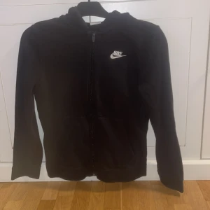 Svart hoodie från Nike med dragkedja - Svart hoodie från Nike med vit logga på bröstet. Tröjan har huva, hel dragkedja framtill och två fickor. Tillverkad i mjukt material, perfekt för chill eller träning. Klassisk och enkel design som funkar året runt.