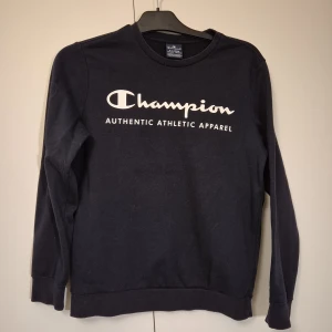Mörkblå Champion sweatshirt 13-14Y/ X-Large - Svart sweatshirt från Champion i storlek 13-14Y/X-Large med vit logga och text på bröstet. Klassisk rund halsringning och långa ärmar. 