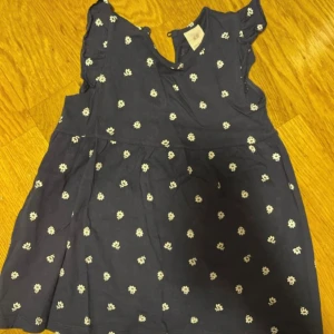 Mörkblå blommig klänning H&Ml  - Supersöt mörkblå kortklänning från H&M med små vita blommor över hela tyget. Klänningen har volangdetaljer vid axlarna och är ärmlös, perfekt för varma dagar. Tillverkad i mjuk bomull som känns skön mot huden.