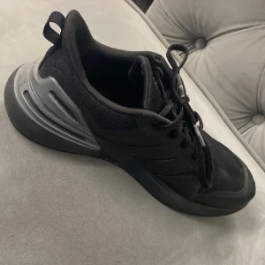 Svarta Adidas sneakers med mesh - Säljer ett par svarta Adidas sneakers med klassiska tre ränder på sidan. Skorna har ovandel i mesh och syntet, snörning och en chunky sula med silvriga detaljer vid hälen. Perfekta för dig som gillar sportig och clean stil.