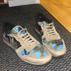 Valentino Rockrunner sneakers camo - Säljer ett par Valentino Rockrunner sneakers med camouflagemönster i blått, grönt och svart. Skorna har detaljer i mocka, skinn och textil samt guld- och vinröda partier vid hälen. Klassisk snörning och platt sula. Perfekt för dig som vill sticka ut med lyxiga och unika sneakers. Sitter bra på mig som brukar ha 45,5 Pris går att diskutera
