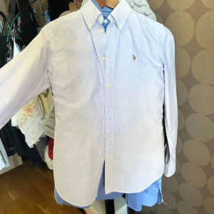 Ljusblå skjorta från Polo Ralph Lauren - Passar xs. Klassisk ljusblå skjorta från Polo Ralph Lauren i bomull med broderad logga på bröstet. Skjortan har button down-krage, långa ärmar och knäppning framtill. Slim-modell som sitter snyggt och passar till många olika stilar.
