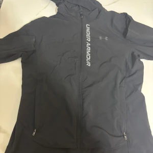 Svart vindjacka från Under Armour - Svart vindjacka från Under Armour med huva och hel dragkedja framtill. Jackan har två sidofickor med dragkedja och logotypen tryckt vertikalt på bröstet. Materialet är lätt och syntetiskt, perfekt för aktiva dagar. Snygg och sportig design med lång ärm.