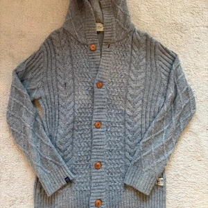 Grå stickad cardigan - Säljer en grå stickad kofta med snyggt flätmönster, stora träknappar och huva. Koftan har långa ärmar och ribbade muddar. Perfekt för lager-på-lager och chill höst- eller vinterstil.