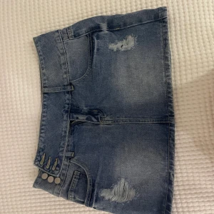 Blå jeanskjol med slitningar - Säljer en snygg blå jeanskjol med slitna detaljer och klassiska fickor både fram och bak. Kjolen har tre silverfärgade knappar i sidan och bälteshällor. Perfekt för dig som gillar en avslappnad och trendig stil. Från shein, köpta för: 250kr?