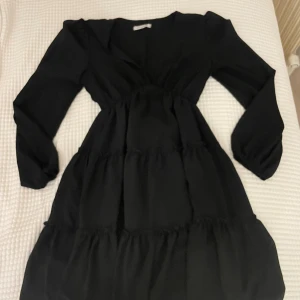 Svart knä långklänning från DesignBySi - Svart knä långklänning från DesignBySi  med v-ringad hals och långa ärmar. Klänningen har volangdetaljer i flera lager och en markerad midja som ger en snygg siluett. Andvänd 1 gång. Köpt för: 699kr 