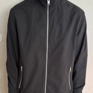 Svart vindjacka från Jack & Jones - Svart vindjacka från Jack & Jones i storlek M. Jackan har hel dragkedja framtill, två fickor med dragkedja och hög krage. (Katt finns i hemmet) nypris 900kr