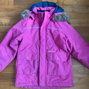 Rosa Didriksons vinterjacka med fuskpäls!!! - Snygg rosa vinterjacka från Didriksons med blått foder och avtagbar huva med fuskpäls. Jackan är 100% vattentät, har stora fickor framtill och reflexdetaljer. Perfekt för kalla dagar och håller dig torr och varm hela vintern.