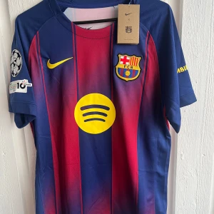 FC Barcelona matchtröja Nike blå/röd - Säljer en FC Barcelona matchtröja från Nike med klassiska blå och röda vertikala ränder, gula detaljer och klubbmärke på bröstet. Tröjan har korta ärmar, Champions League-märke på ärmen och stor gul 27:a på ryggen. Materialet är lätt och andas, perfekt för fotboll.